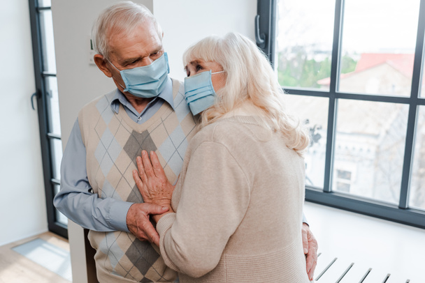 sad elderly couple in medical masks hugging at home during self isolation - 写真・画像