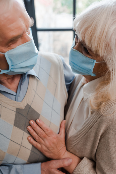happy elderly couple in medical masks hugging at home during quarantine - Fotó, kép