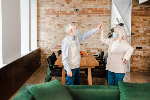 feliz pareja de ancianos en máscaras médicas bailando en casa durante el auto aislamiento
 - Foto, Imagen