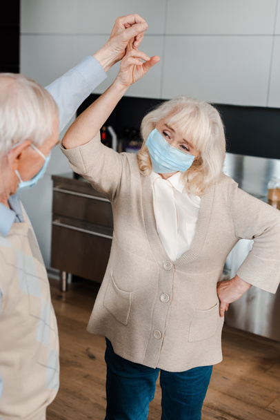 happy elderly couple in medical masks dancing at home during self isolation - Fotografie, Obrázek