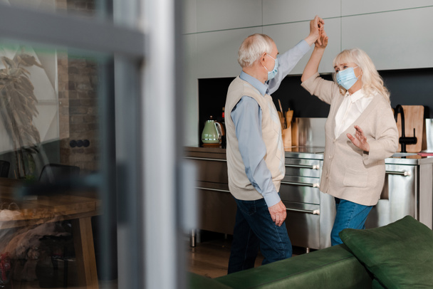 elderly couple in medical masks dancing on kitchen during self isolation - Foto, imagen