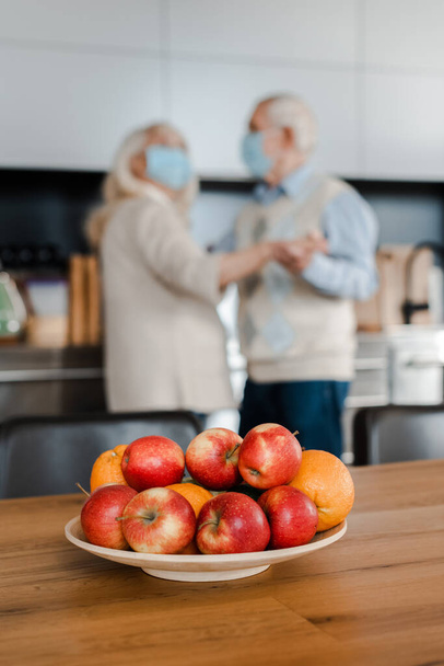 selective focus of elderly couple in medical masks dancing on kitchen with fruits during quarantine - Фото, изображение