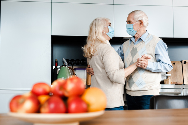 ηλικιωμένο ζευγάρι με ιατρικές μάσκες χορεύουν στην κουζίνα με μήλα κατά τη διάρκεια της αυτο-απομόνωσης, επιλεκτική εστίαση - Φωτογραφία, εικόνα