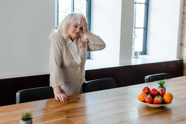 tired elderly woman with headache standing at table with fruits during quarantine - Photo, Image