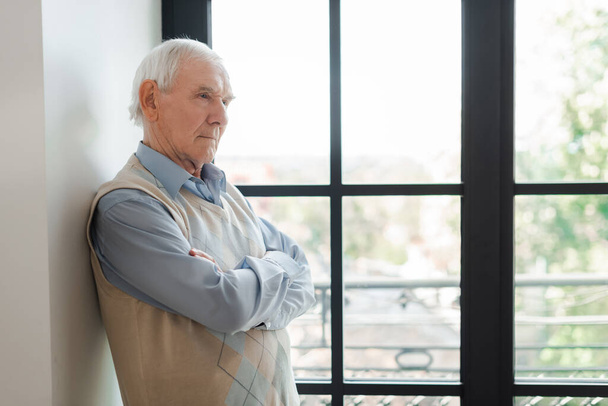 starszy samotny mężczyzna z skrzyżowanymi ramionami stojący przy oknie podczas samoizolacji - Zdjęcie, obraz