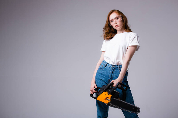 Sexy redhead girl in safety glasses in a white T-shirt and jeans holds a chainsaw and looks at the camera - Foto, Imagen
