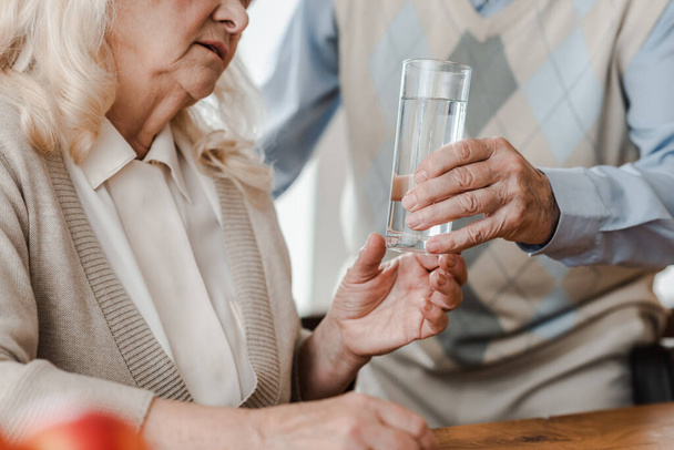 cropped view of elderly husband and ill wife with glass of water at home during quarantine - Foto, afbeelding