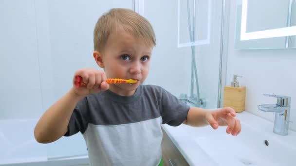 幸せな少年は歯をブラッシングしている。笑顔の子供は小さな椅子に立っている間、軽い白いバスルームで彼の両親に彼の噛んだ歯を示しています。子供のための水活動、衛生、ケア - 映像、動画