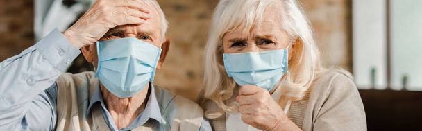 worried elderly couple in medical masks sitting at home during self isolation, horizontal concept - Photo, Image