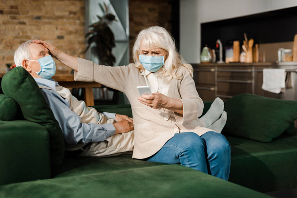 mujer anciana preocupada en máscara médica llamando al médico con teléfono inteligente mientras está enfermo marido con fiebre acostado en el sofá durante la epidemia de coronavirus
  - Foto, Imagen
