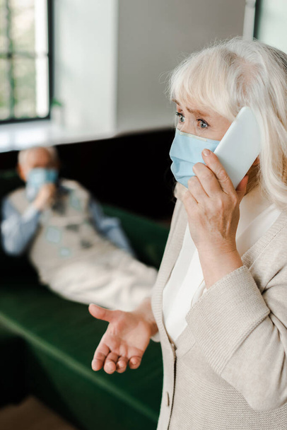 worried elderly wife in medical mask calling doctor with smartphone while ill husband lying on sofa during coronavirus epidemic, selective focus - Foto, imagen