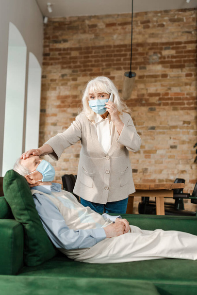 elderly wife in medical mask calling doctor with smartphone while ill husband with fever lying on sofa during self isolation - Фото, изображение