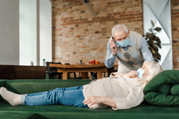 Älterer Mann mit medizinischer Maske spricht auf Smartphone, während kranke Frau während Selbstisolierung auf Sofa liegt - Foto, Bild