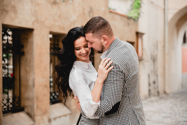 Charming girl with long brown hair and a man with a beard kissing on the background of the morning city. Newlyweds on their wedding day - Photo, image