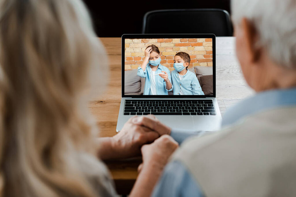 grandparents holding hands and having video chat with grandchildren in medical masks holding thermometer during self isolation - Foto, Imagem