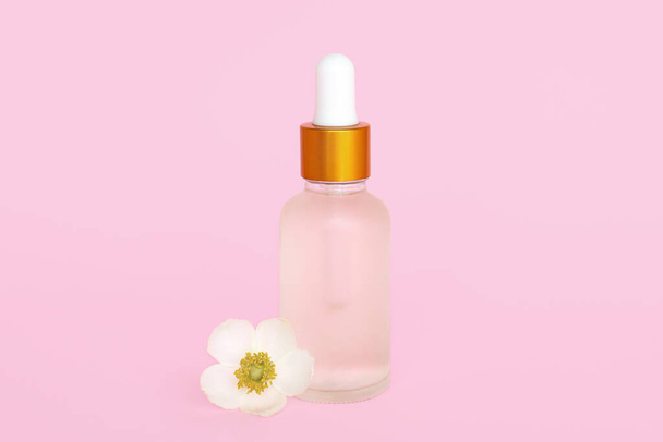 Bouteille cosmétique en verre avec huile. Conteneur en verre pour un produit cosmétique pour les femmes avec de petites fleurs blanches sur fond rose
. - Photo, image