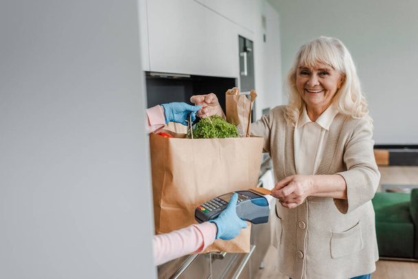 onnellinen iäkäs nainen ottaa ruoan toimitus ja maksaa luottokortilla ja terminaali aikana coronavirus pandemia - Valokuva, kuva