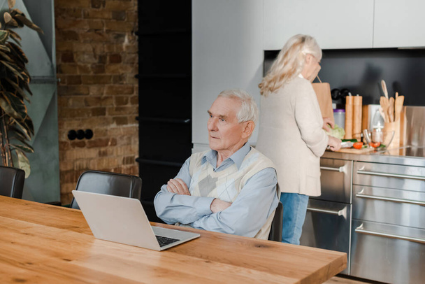 marido anciano pensativo usando el ordenador portátil mientras la esposa cocina en la cocina durante la cuarentena
 - Foto, Imagen