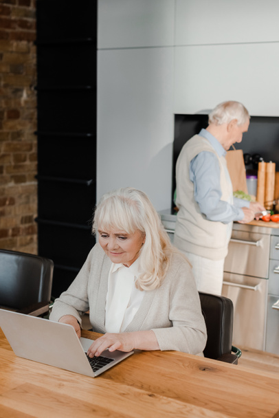 moglie anziana utilizzando il computer portatile mentre il marito cucina in cucina durante l'isolamento
  - Foto, immagini