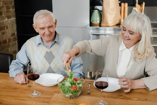 smiling senior couple having dinner with red wine and salad at home on quarantine - Fotó, kép