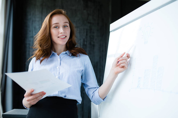 Attractive smiling girl in a blue shirt and skirt conducts business training on a white board and looks at the camera - Foto, Bild
