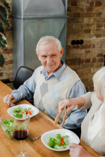 positief bejaard paar met diner met rode wijn en salade thuis op quarantaine - Foto, afbeelding