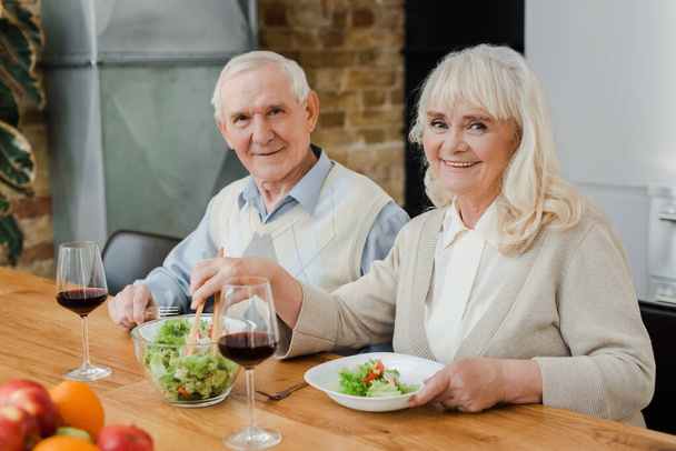 smiling senior couple having dinner with wine and salad at home on self isolation - Foto, Bild