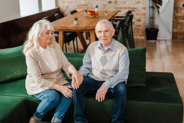 расстроенная пожилая пара, держащаяся за руки на диване во время самоизоляции
 - Фото, изображение