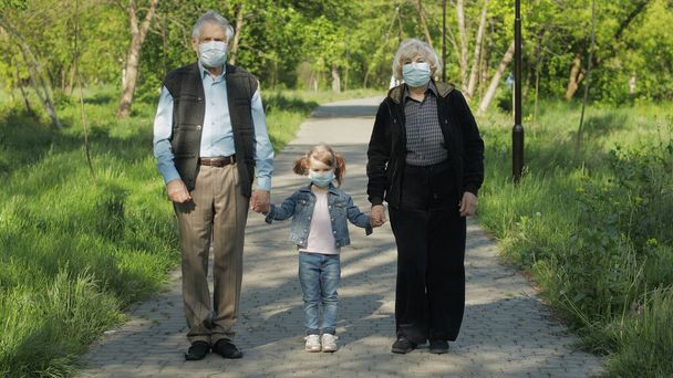 医療マスクの孫娘を持つ古い祖父母は公園を歩く。コロナウイルス - 写真・画像