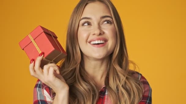 Young cheery girl isolated over yellow wall background holding present box trying to guess what's inside - Metraje, vídeo