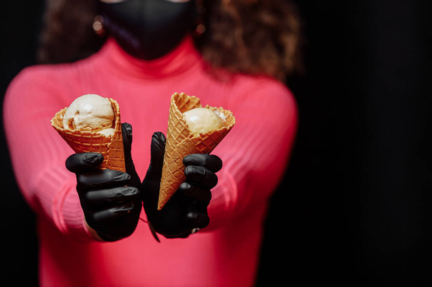 黒の保護手袋の手はアイスクリームとワッフルコーンを保持しています。コロノウイルスからの保護。隔離中にアイスクリームを販売する概念。保護黒手袋で手にアイスクリーム. - 写真・画像