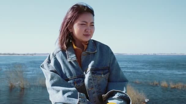 Young asian woman in a jeans jacket coughing while sitting on the river bank - Imágenes, Vídeo
