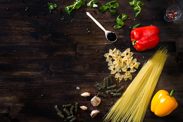 Various sorts of pasta on top of a dark wooden background. Farfalle, fusilli, spaghetti, bell pepper, corn salad, garlic cloves, red, green or yellow pepper. - Photo, image