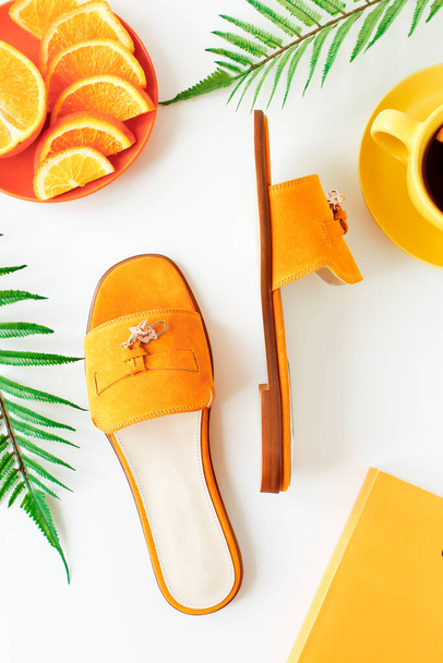 Yellow stylish pair of clog shoes, cup with tea and orange slice, green palm leaves on a white background. Flat lay, top view - Foto, immagini