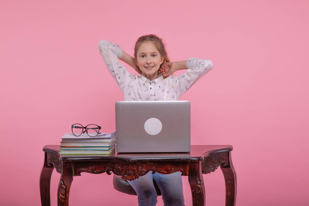 Gyönyörű fiatal lány kiegyenesíti hosszú szőke haját, miközben egy számítógép előtt ül. Egy halom könyv egy laptop mellett. Rózsaszín alapon. - Fotó, kép