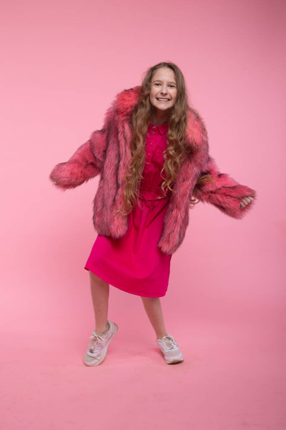 A cheerful girl in fashionable clothes, a light pink fur coat, a pink bright dress and white sneakers is dancing on a pink background. - 写真・画像
