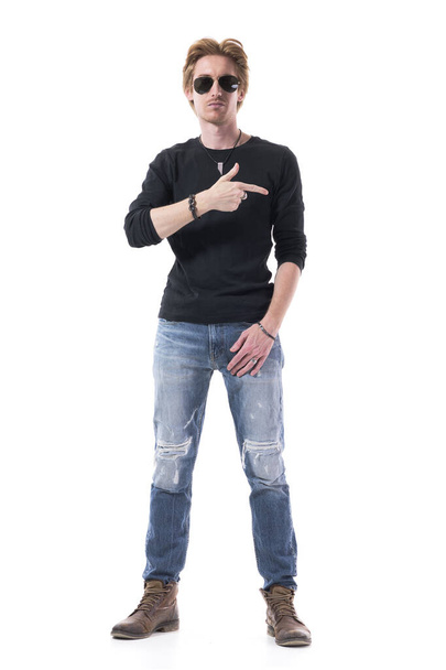 Ernstige houding stijlvolle jonge man met een zonnebril dragen jeans wijzen naar kant beschuldigen of beschuldigen. Volledige lichaamslengte geïsoleerd op witte achtergrond.  - Foto, afbeelding