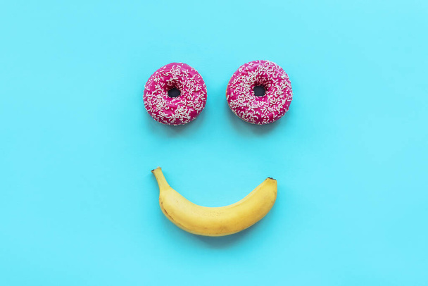 Dos rosquillas y un plátano en forma de una cara sonriente sobre fondo azul
 - Foto, Imagen