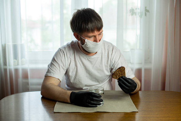 COVID-19. Young caucasian man in glasses, white mask and black gloves with a slice of bread and a cup of water in his hands in the kitchen during a pandemic. Coronavirus in Europe. - 写真・画像