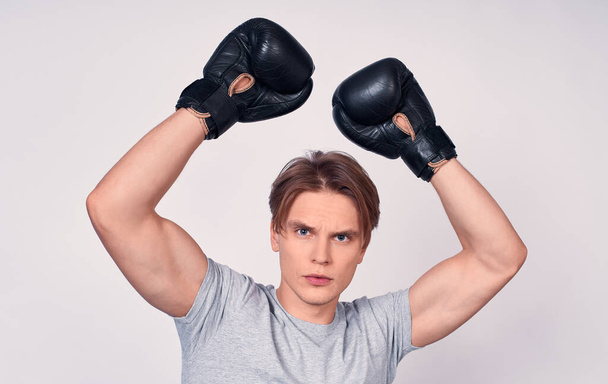 Young handsome Caucasian sports youth, with blond hair, in a gray basic t-shirt, with black boxing gloves celebrates his victory, raised his hands up, looks straight. - Valokuva, kuva
