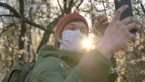 o homem em uma máscara médica toma uma selfie no telefone com uma macieira florescente ao pôr-do-sol
 - Filmagem, Vídeo