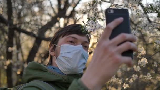 Coronavirus karantinası sırasında selfie, tıbbi maske takmış bir adam günbatımında akıllı telefonda çiçek açan elma ağacıyla fotoğraf çekiyor. - Video, Çekim