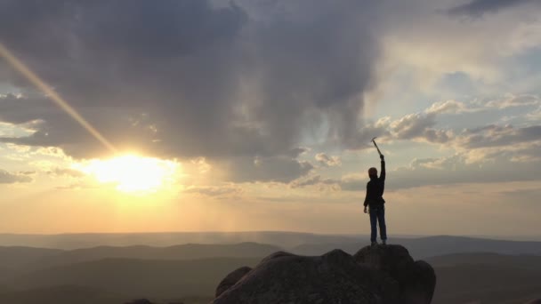 Drone shot van een silhouet van een bergbeklimmer die op de top van een berg staat en overwinnend zijn hand optilt met een ijsbijl bij zonsondergang. - Video