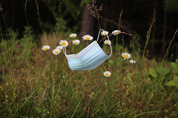 Surgical mask hangs on camomile flowers in summer meadow. Virus protection. Equipment for protection from coronavirus - Φωτογραφία, εικόνα