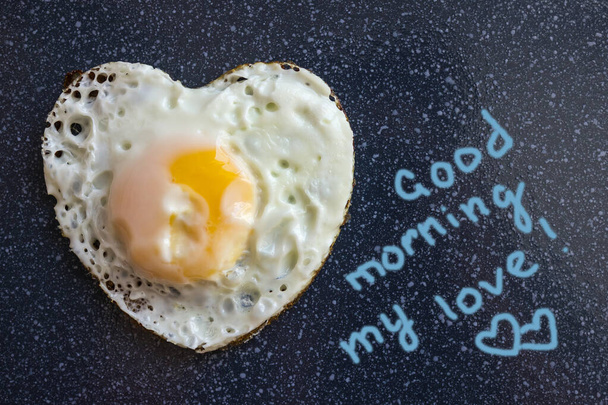 Ein einfaches Frühstück, mit Liebe zubereitet. Gebratenes Ei in Herzform. Ein guter Start in den Tag. Dunkler Hintergrund, Ansicht von oben. Die Inschrift stammt vom Autor. - Foto, Bild