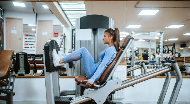 Une jolie jeune fille caucasienne en équipement de sport bleu sur une machine à jambes dans la salle de gym
 - Photo, image
