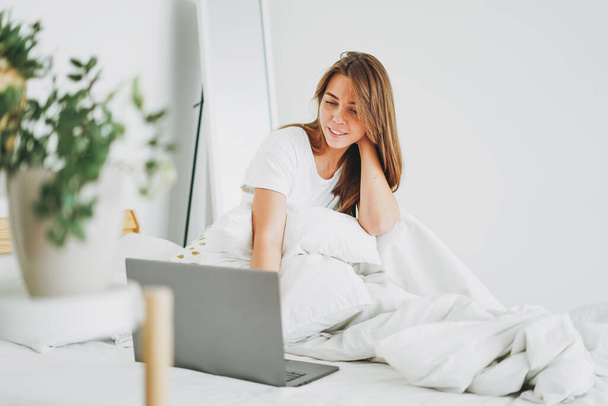 Jeune femme brune pigiste en vêtements décontractés avec ordinateur portable travaillant dans le lit à la maison dans un séjour intérieur lumineux à la maison
 - Photo, image