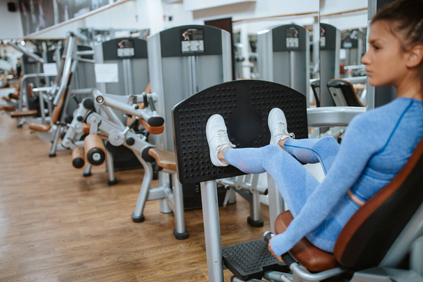 Atrakcyjna młoda biała dziewczyna w niebieskim sprzęcie sportowym na maszynie do nóg w siłowni - Zdjęcie, obraz