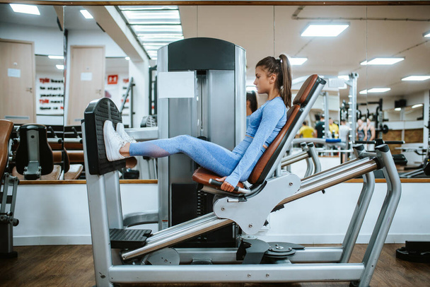 Uma jovem menina caucasiana atraente em equipamento esportivo azul em uma máquina de perna no ginásio
 - Foto, Imagem