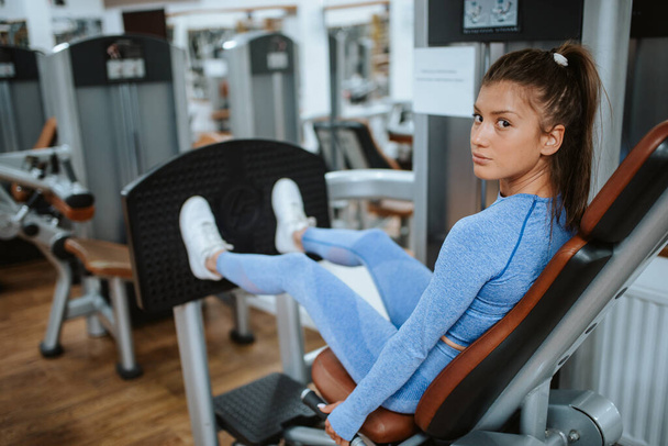 An attractive young caucasian girl in blue sports equipment on a leg machine in the gym - Photo, Image